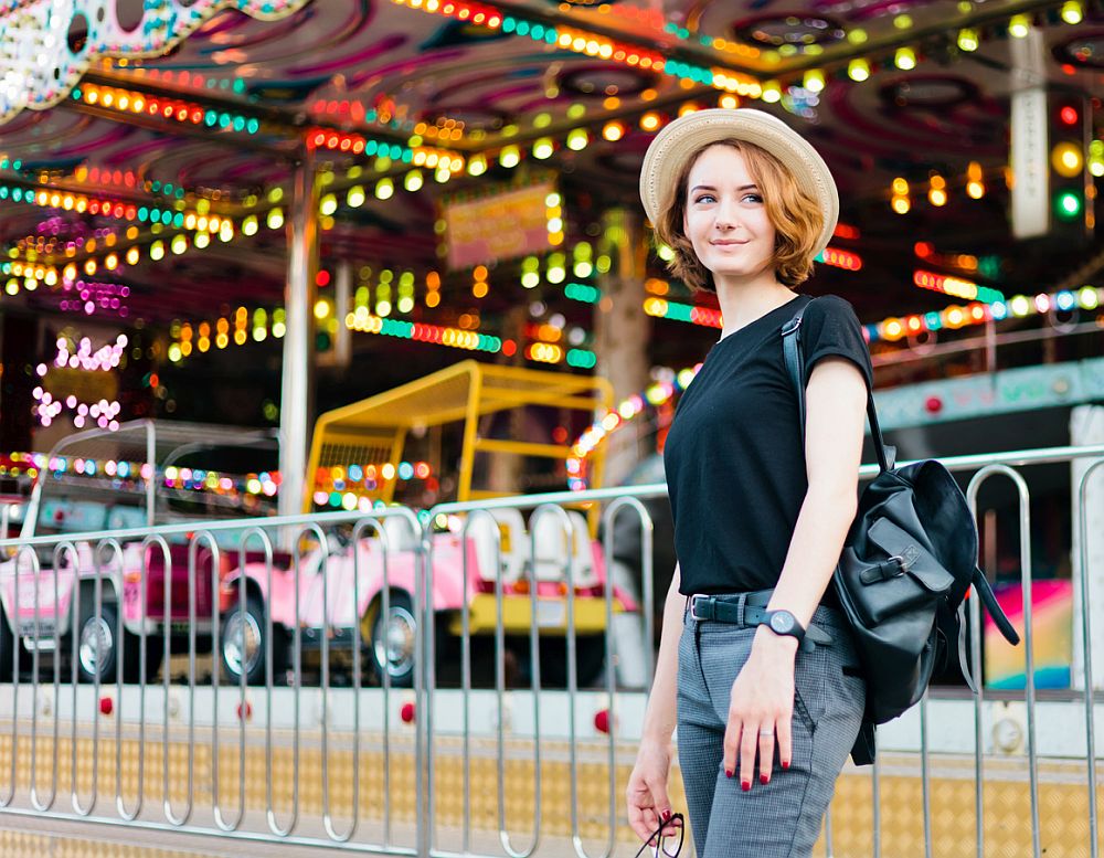 Young cheerful hipster woman in glasses and hat in amusement park
