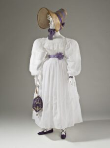 A mannequin dressed in white, with a purple reticule on hand.